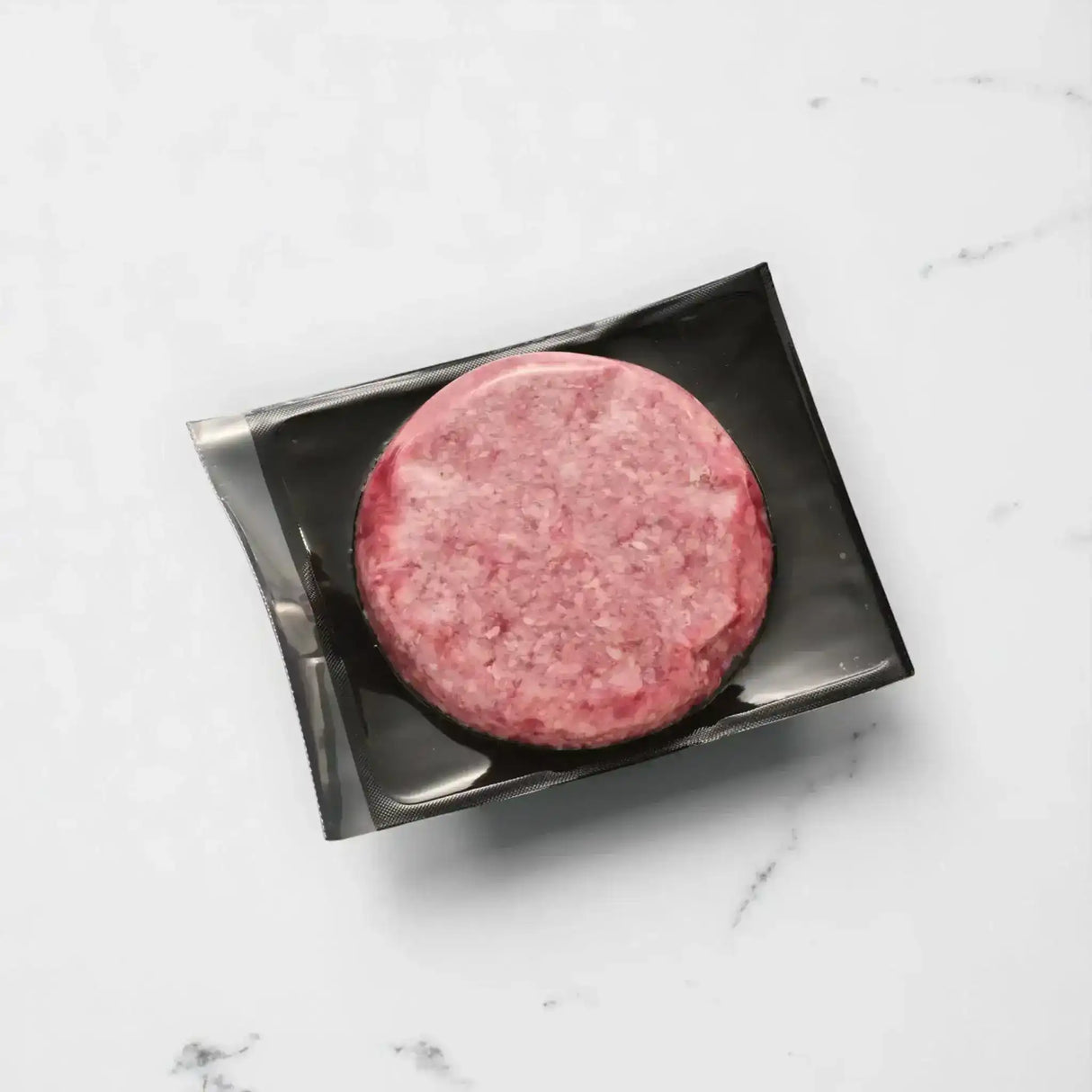 Vacuum-sealed raw beef burger patty on black tray, marble surface background