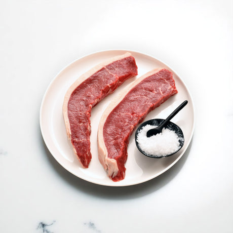 Two raw Grain fed picanha steaks on a white plate with a bowl of coarse salt