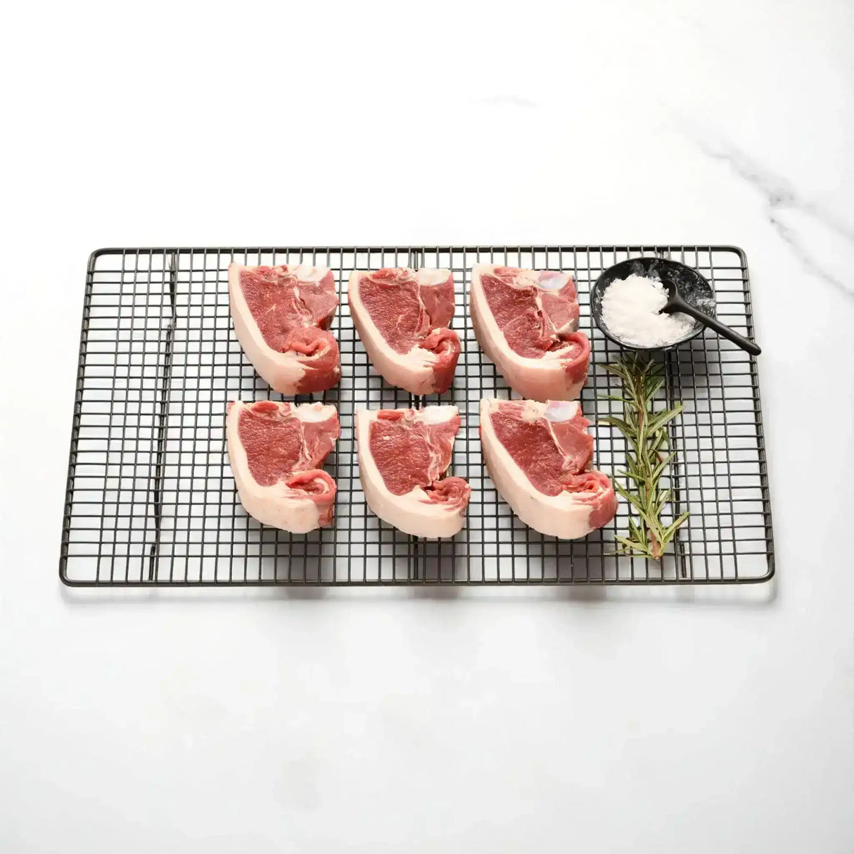 Six fresh lamb loin chops on a wire rack with rosemary and a small bowl of sea salt
