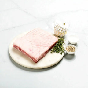 Fresh pork belly slab on a round board with herbs, white peppercorns, and salt from Peter Augustus