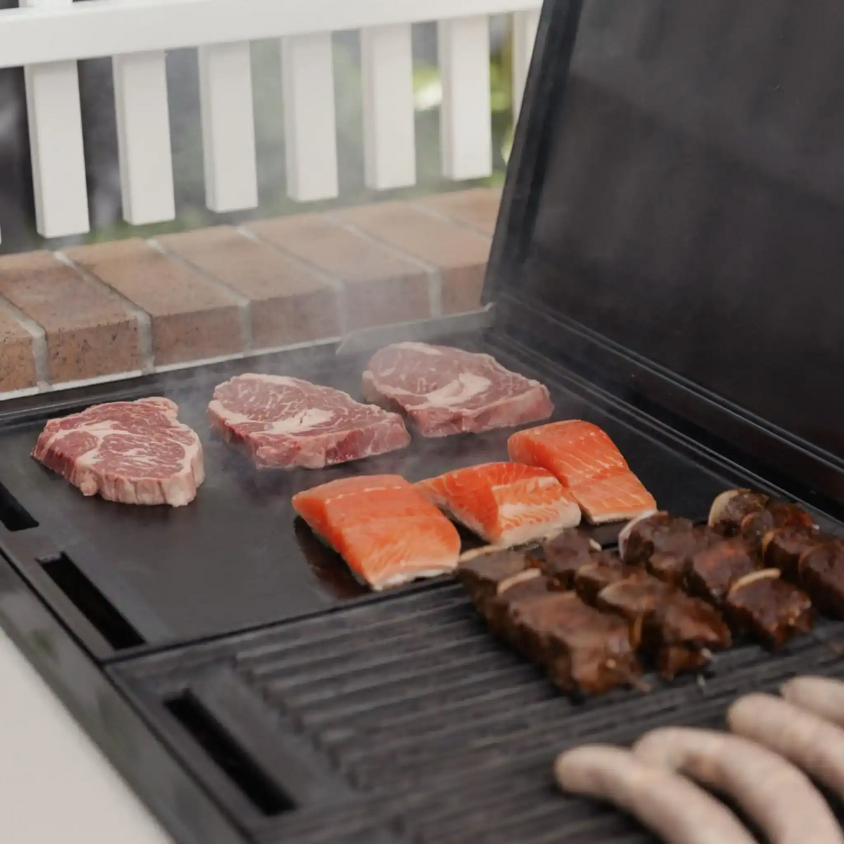 Assorted beef steaks, salmon fillets, skewers, and sausages grilling on a barbecue.