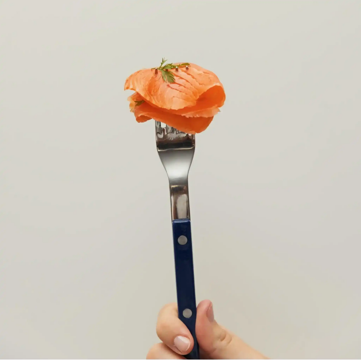 Hand holding fork with sliced smoked salmon garnished with dill against white background