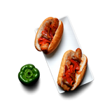 Two hot dogs in white buns topped with red peppers and onions, arranged on a white rectangular plate with a whole green bell pepper on a white background.