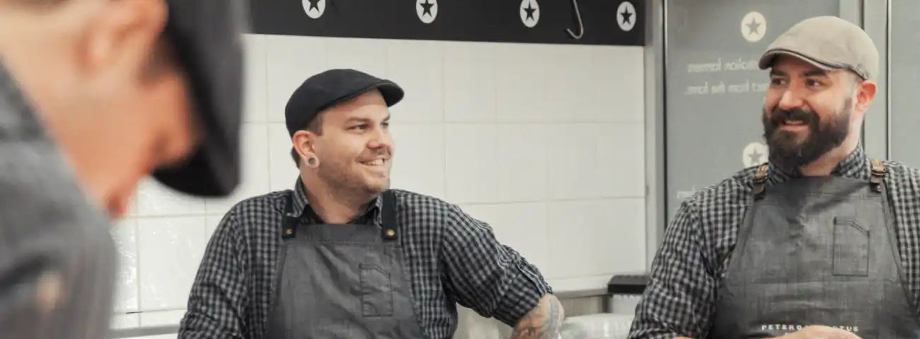 Two men in a kitchen setting, one wearing a cap and apron, the other in a blurred foreground.