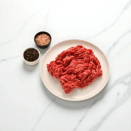 Fresh Wagyu beef mince on a white plate with bowls of peppercorns and pink salt on marble