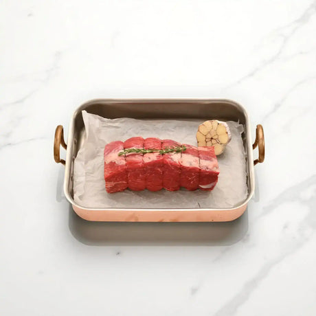 Raw wagyu rump roast tied with twine and rosemary, with garlic in a roasting pan on marble