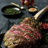 Sliced Wagyu Tomahawk steak topped with herbs and seasoning on a dark serving board.