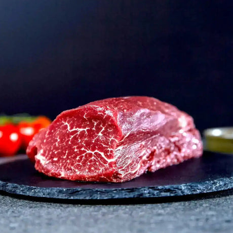 Marbled raw beef tenderloin chateaubriand cut on slate platter with tomatoes and oil in background