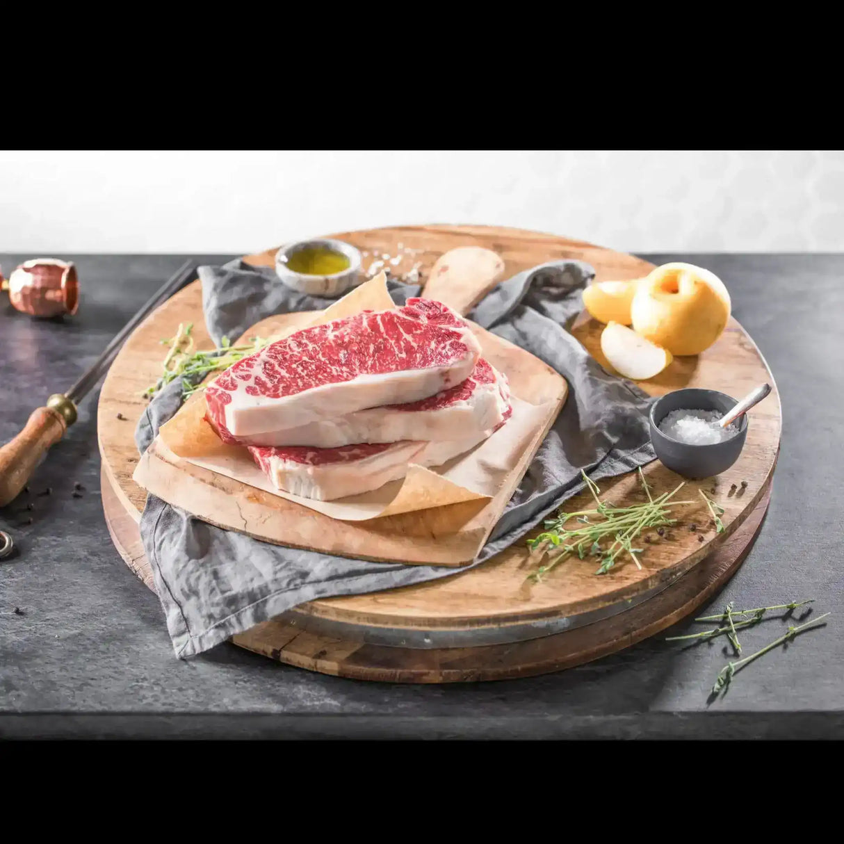 Raw Wagyu striploin steak with heavy marbling on a wooden board, surrounded by salt, herbs, and oil.